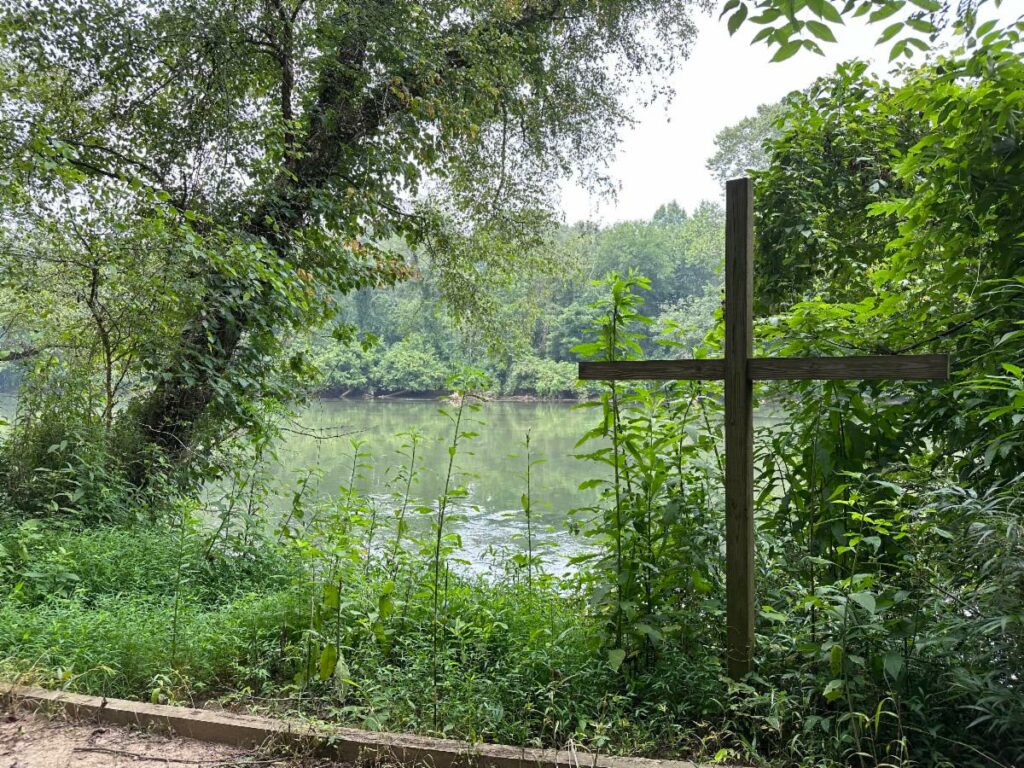 A beautiful scenery of a lake between shrubs and trees, with a wooden cross standing on the right hand side